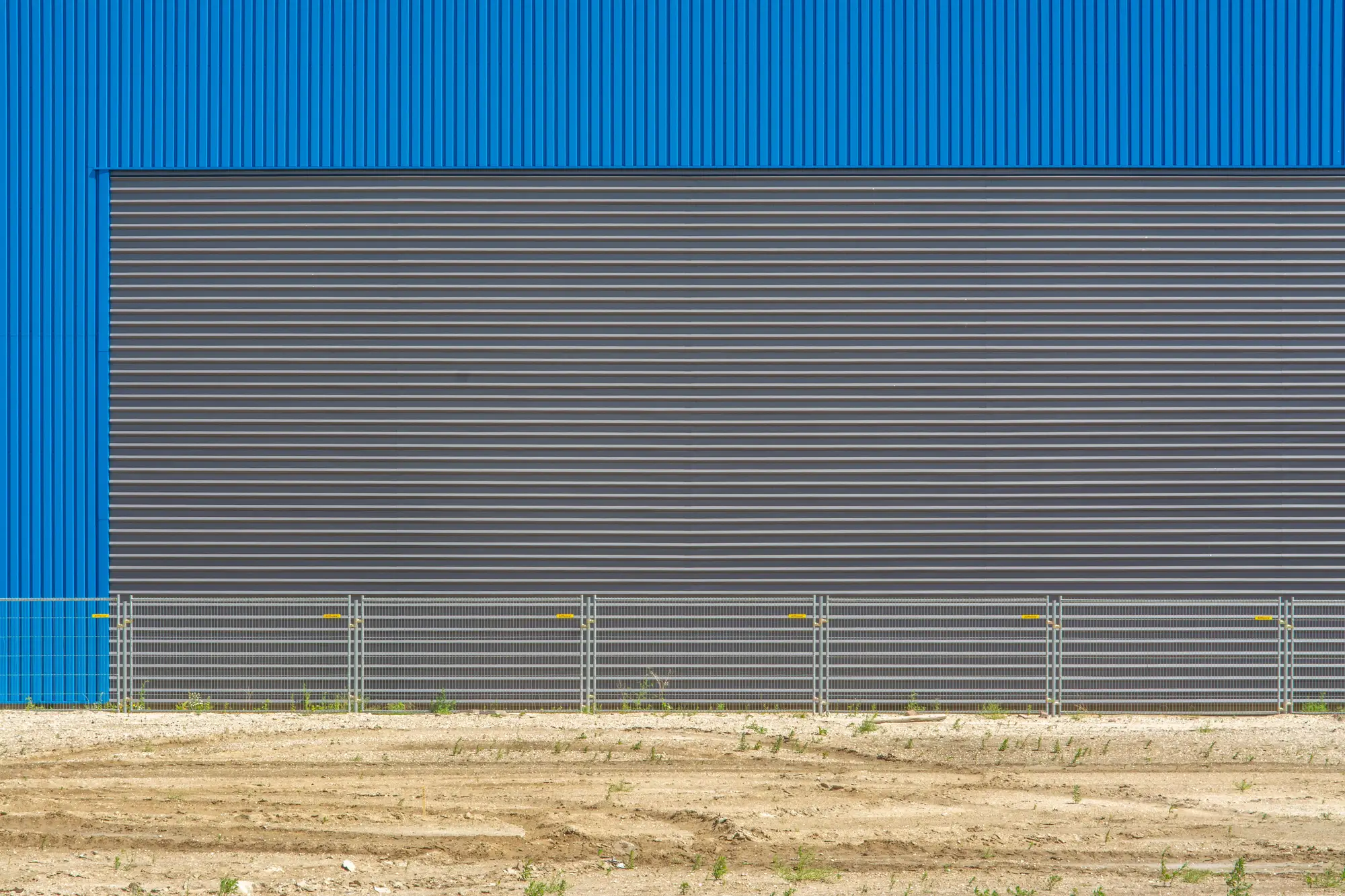 Corrugated wall of a newly built warehouse behind a site fence, with graded bare ground in front